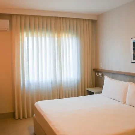 Costao Do Santinho Resort All Inclusive Florianópolis Habitación foto This is an image of a bright and modern hotel or guest room.

**Key elements in the photo:**

*   **Bed:** A large, comfortable-looking bed dominates the foreground, neatly made with crisp white linens.
*   **Window Treatment:** A large window covers